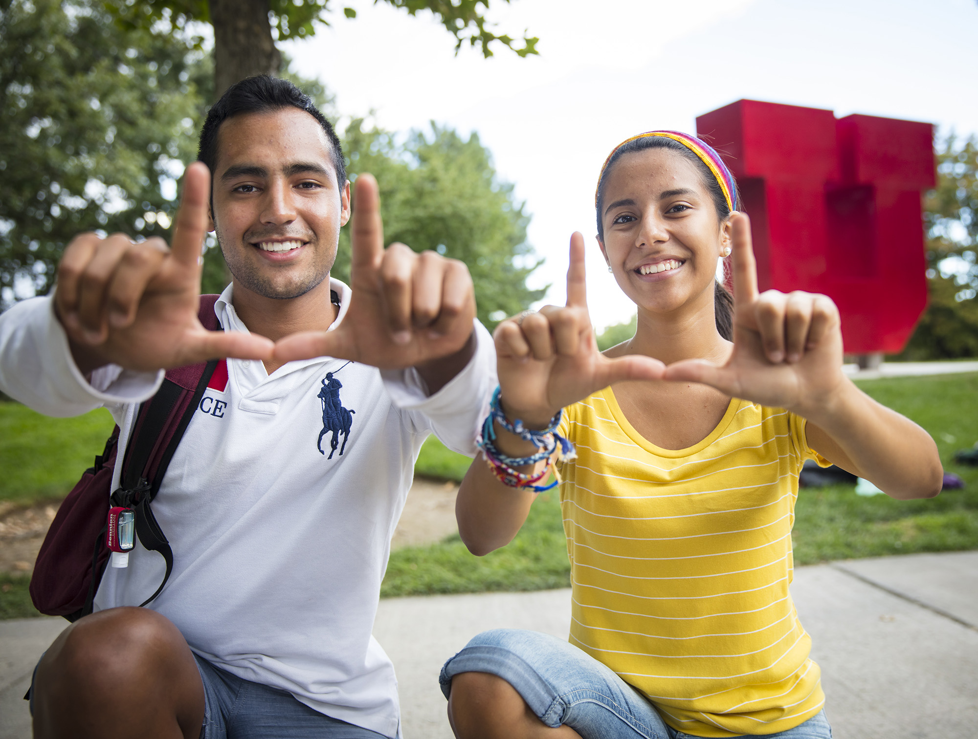2 students sitting in front of the large red U sculpture making a "U" with their hands