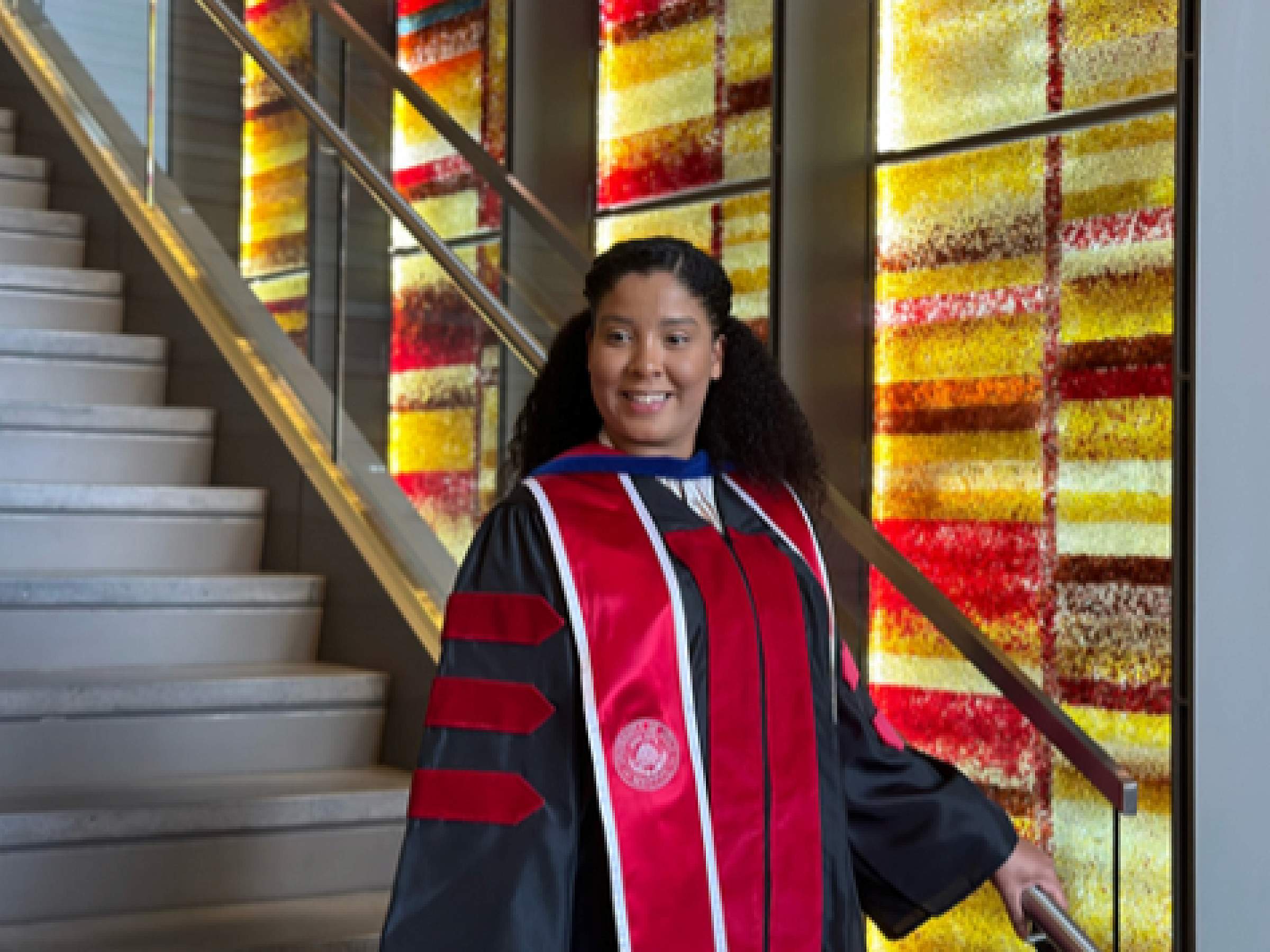 Marysol Almestica-Roberts, PhD, poses in Convocation attire on steps inside the University of Utah College of Pharmacy atrium.