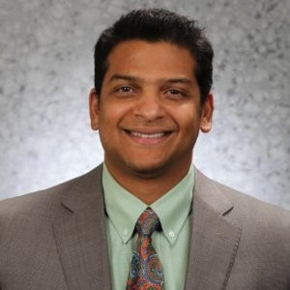Dr. Venkata Kashyap Yellepeddi smiles in portrait photo with grey suit, green shirt, and orange tie