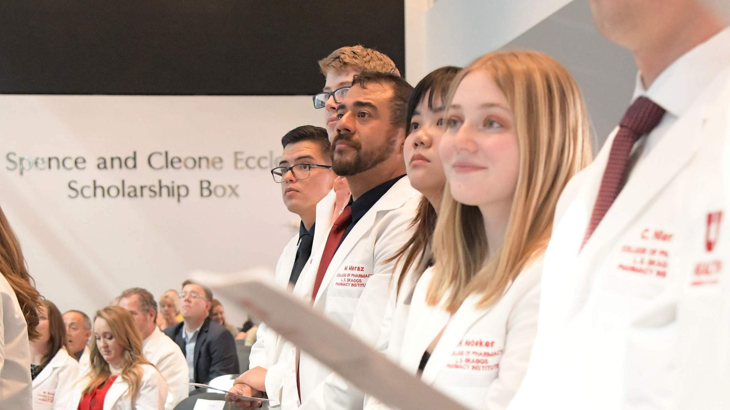 Students stand and hold their program, listening intently, at the 2023 College of Pharmacy White Coat Ceremony.